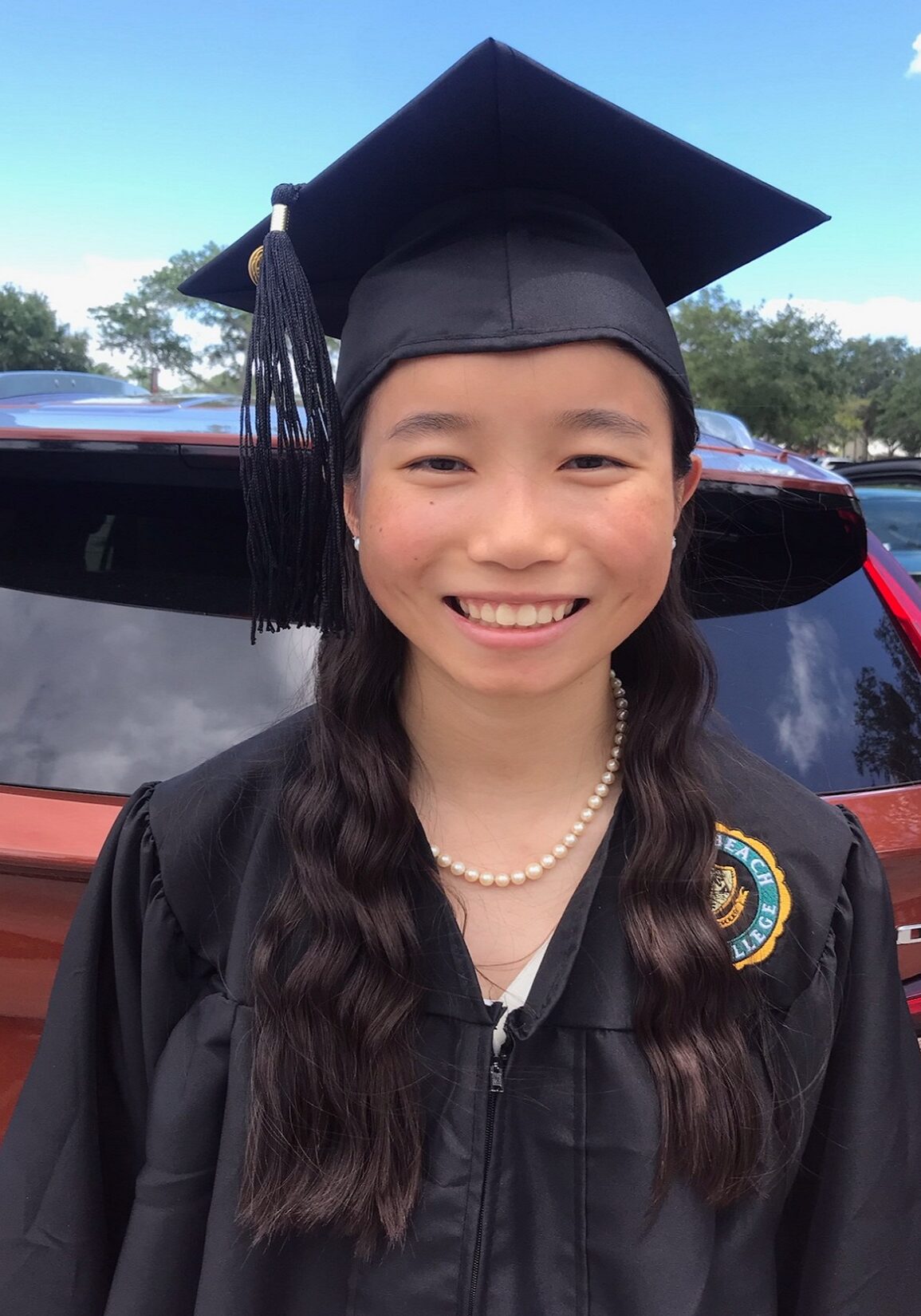 A girl in graduation attire standing next to a red car.