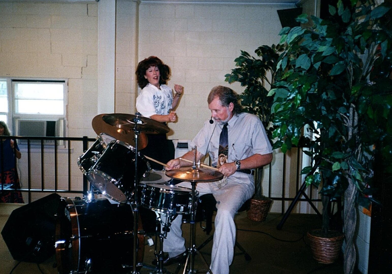 A man and woman playing drums in front of plants.